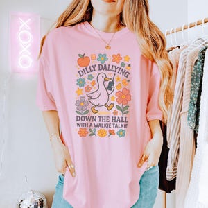 May include: Pale pink t-shirt with a cartoon duck holding a walkie-talkie, surrounded by flowers and the text "Dilly Dallying Down the Hall with a Walkie Talkie."