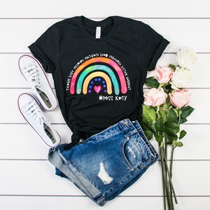 May include: A black short-sleeved t-shirt featuring a colourful rainbow graphic with the text Teach Love Inspire Motivate Lead Encourage Listen Connect and #MISS KATY, paired with blue denim shorts and white canvas trainers.