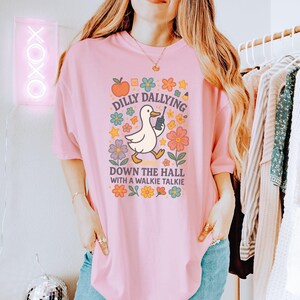 May include: Pale pink t-shirt with a retro graphic. The design features a cartoon duck with a walkie-talkie, surrounded by flowers, and the text "Dilly Dallying Down the Hall with a Walkie Talkie." The style is whimsical.