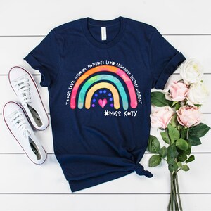 May include: A navy blue short-sleeve t-shirt featuring a colourful rainbow graphic with the text "TEACH LOVE INSPIRE MOTIVATE LEAD ENCOURAGE LISTEN CONNECT" and "#MISS KATY" printed in white, accompanied by a small pink heart and blue dots.