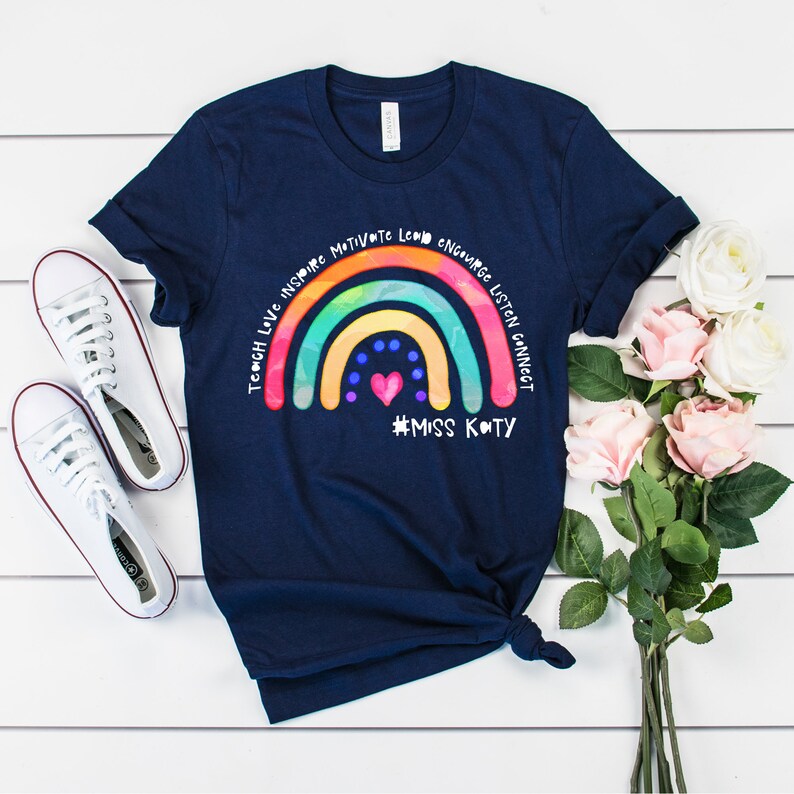 May include: A navy blue short-sleeve t-shirt featuring a colourful rainbow graphic with the text "TEACH LOVE INSPIRE MOTIVATE LEAD ENCOURAGE LISTEN CONNECT" and "#MISS KATY" printed in white, accompanied by a small pink heart and blue dots.
