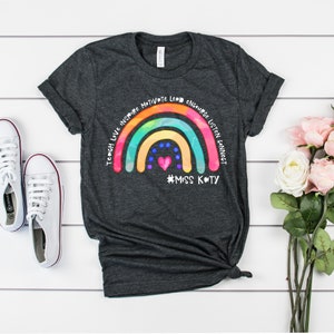 May include: A dark heather grey t-shirt featuring a colourful rainbow graphic with the words Teach, Love, Inspire, Motivate, Lead, Encourage, Listen, Connect arched above it. A small pink heart and blue dots sit below the rainbow with #MISS KATY.