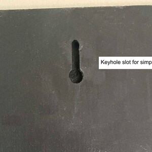 May include: A close-up of a black surface with a keyhole slot for simple mounting. The keyhole slot is a common feature for hanging items on a wall.