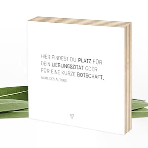 Könnte beinhalten: Ein quadratischer Holzblock mit einer weißen Vorderseite, bedruckt mit dem schwarzen Text: HIER FINDEST DU PLATZ FÜR DEIN LIEBLINGSZITAT ODER FÜR EINE KURZE BOTSCHAFT. NAME DES AUTORS. Ein kleines schwarzes Herzsymbol ist unten in der Mitte aufgedruckt.