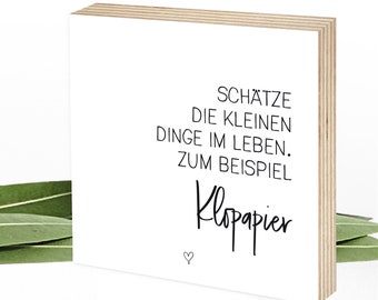 May include: White wooden sign with black text that reads "Schätze die kleinen Dinge im Leben. Zum Beispiel Klopapier".