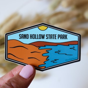 Op de afbeelding: Een blauwe en oranje sticker met de tekst "SAND HOLLOW STATE PARK" en een afbeelding van een woestijnlandschap met een meer.