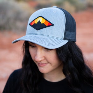 May include: A gray and black trucker hat with a patch featuring a mountain range and a setting sun. The patch is in shades of orange, yellow, and red.