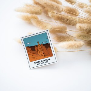 May include: A white sticker with a cartoon illustration of a red rock formation in Bryce Canyon National Park. The sticker has the text "Bryce Canyon National Park" printed on it.