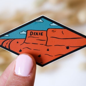 May include: A diamond-shaped sticker with a cartoon illustration of a red rock formation with the word "DIXIE" written on it. The background is a blue sky with white clouds.