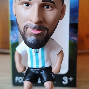 May include: A figurine of a soccer player wearing a white and light blue jersey with the number 10 on it. The player is kicking a white and black soccer ball. The figurine is on a black base.