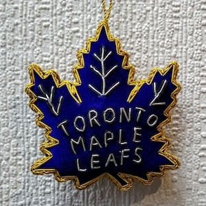 Może przedstawiać: Niebieska ozdoba w kształcie liścia klonu ze złotym obramowaniem i srebrnym napisem "TORONTO MAPLE LEAFS". Zawieszona na złotym sznurku. Tło to teksturowana biała powierzchnia.