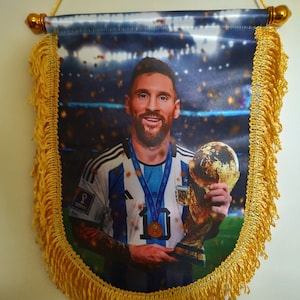 Puede incluir: Un banderín decorativo con un retrato de un futbolista sosteniendo un trofeo. El banderín tiene flecos dorados y un cordón dorado para colgar. El jugador lleva una camiseta a rayas azules y blancas.