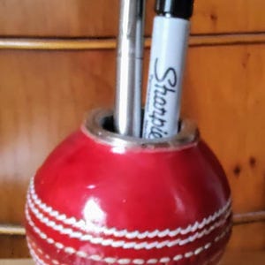 May include: A red cricket ball-shaped pen holder with white stitching, holding a silver pen and a black Sharpie marker. The holder sits on a small wooden block.