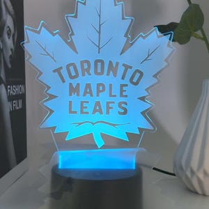 Puede incluir: Una luz nocturna impresa en 3D azul y blanca con forma de hoja de arce con las palabras "Toronto Maple Leafs" impresas en ella. La luz está sobre una base negra.