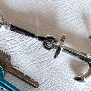 Puede incluir: Un llavero de ancla plateada con una correa de cuero trenzado marrón y un llavero plateado. Se ven dos llaves con cabezas azules y plateadas. El llavero está sobre una superficie blanca texturizada.