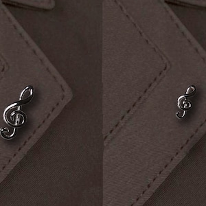 Puede incluir: Dos pines de solapa de clave de sol plateados sobre un fondo de tela marrón. El pin de la izquierda es más grande que el de la derecha.