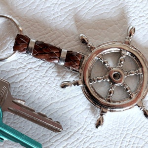 Puede incluir: Un llavero plateado con forma de timón de barco, con un mango de cuero marrón y un llavero de metal. Se ven dos llaves, una turquesa y otra plateada. El llavero está sobre una superficie blanca texturizada.