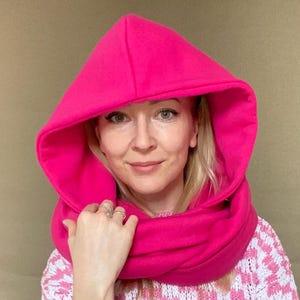 Könnte beinhalten: Ein leuchtend pinkfarbener Fleece-Kapuzen-Schal mit einem langen, breiten Hals. Der Schal wird von einer Person getragen, die einen pink-weiß karierten Pullover und eine weiße Hose trägt.
