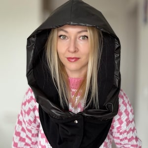 Puede incluir: Un pañuelo con capucha negro con un exterior brillante similar al cuero y un interior mate y suave. El pañuelo se abrocha en la parte delantera y se lleva sobre un suéter con estampado rosa y blanco. La capucha está diseñada para proteger del frío.