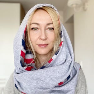 Puede incluir: Bufanda con capucha gris con un patrón de rayas rojas, blancas y azules en el interior. La bufanda está envuelta alrededor de la cabeza y el cuello, creando un look acogedor y elegante. La persona lleva un suéter gris.
