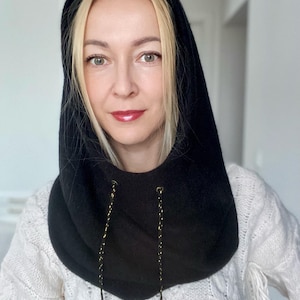 Könnte beinhalten: Ein schwarzer Kapuzenschal mit goldfarbenen Kordeln. Der Schal ist aus einem weichen, dunklen Stoff gefertigt und wird um Hals und Kopf getragen. Die Person auf dem Bild trägt einen weißen Pullover.