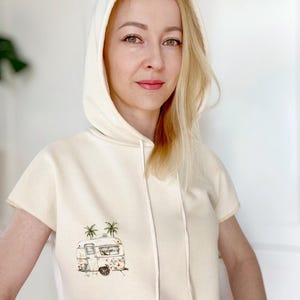 Pode incluir: Uma sudadera com capuz de manga curta de cor creme com cordão de ajuste. A sudadera apresenta uma ilustração de estilo vintage de uma autocaravana com palmeiras em fundo.