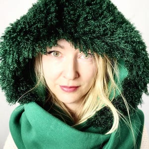 May include: A green hooded scarf with a fuzzy texture. The scarf is wrapped around a person's neck and the hood is pulled up over their head.
