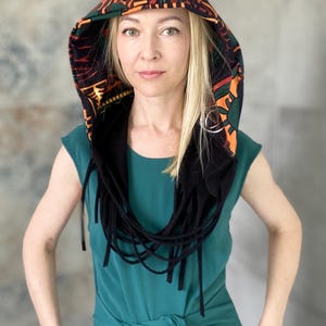 Könnte beinhalten: Ein Kapuzenschal mit schwarzer Basis und einem farbenfrohen geometrischen Muster in Orange, Grün und Braun. Der Schal hat schwarze Fransen. Die Person trägt ein türkisfarbenes Kleid.