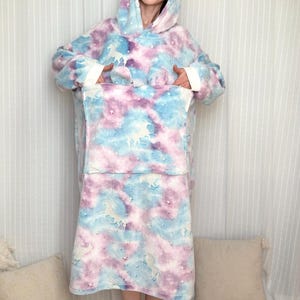 Pode incluir: Um cobertor com capuz oversized com um padrão tie-dye em azul, roxo e branco. Possui um grande bolso frontal e punhos brancos. O design inclui um padrão de unicórnio.