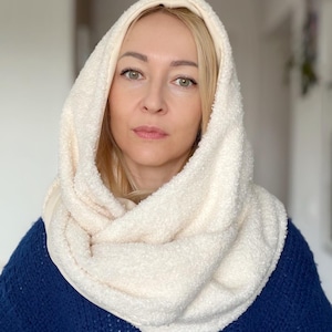 May include: A cream-coloured, hooded scarf made of a soft, textured material. The scarf is wrapped around the neck and head, creating a cosy and warm look. The person is wearing a blue jumper.