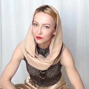 Può includere: Una donna indossa un copricapo color beige e un top senza maniche con un motivo leopardato. Il top è abbinato a pantaloni dorati. La donna ha i capelli biondi e rossetto rosso.