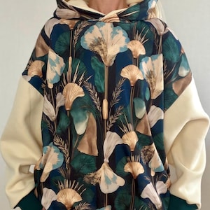 Può includere: Una felpa con cappuccio a maniche lunghe caratterizzata da uno sfondo blu scuro con un motivo ripetuto di fiori a ventaglio ed erbe alte nei colori bianco, oro e verde. Le maniche e l'orlo sono color crema, con polsini verde scuro.