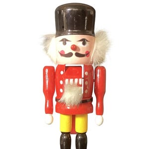 May include: A wooden nutcracker figurine in a soldier's uniform. The figurine has a black hat, a red jacket with white buttons, yellow pants, and black boots. It stands on a green base and has white hair and a mustache.