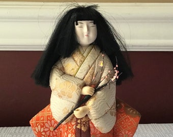 May include: A traditional Japanese doll wearing a white and orange kimono with gold accents. The doll has black hair and a painted porcelain face. The doll is holding a branch with pink blossoms.