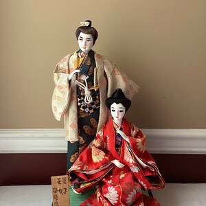 May include: A pair of traditional Japanese dolls dressed in elaborate costumes. The male doll is wearing a green and gold robe with a sword, while the female doll is wearing a red and gold robe with floral patterns. The dolls are sitting on a black base with the text "流 琳 人形" in Japanese.