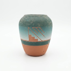 May include: A small, handcrafted ceramic vase with a rounded body. The vase features a gradient of teal and green hues, with a terracotta base and a geometric design in orange. The design includes a stepped pattern and angular lines.