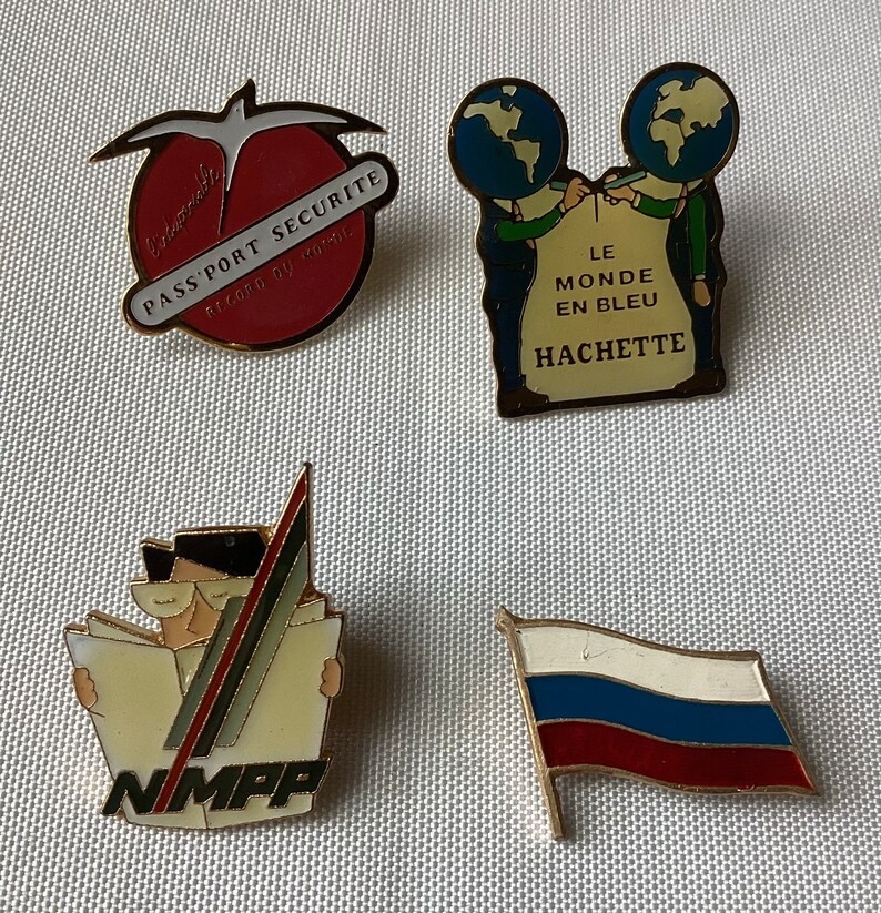Op de afbeelding: Vier emaillepins met verschillende ontwerpen. De eerste pin is rood met een witte vogel en de tekst "Universitaire Pass'Port Securite Record du monde". De tweede pin is een blauwe en groene wereld met twee figuren die een steen vasthouden met de tekst "Le Monde en Bleu Hachette". De derde pin is geel met een figuur die een bril en een hoed draagt en een kaart vasthoudt met de tekst "NMPP". De vierde pin is een rood, wit en blauwe vlag met een gouden paal.