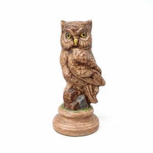 May include: A ceramic owl figurine featuring brown and tan feathers with detailed texture. The owl has bright yellow eyes and a yellow beak, perched on a brown rock base that sits atop a circular, wood-toned pedestal with a smooth finish.