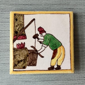 May include: A hand-painted ceramic tile depicting a person in a green shirt and yellow pants using a hammer to work on a metal object in a forge. The tile has a yellow border.