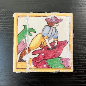 May include: A hand-painted ceramic tile with a yellow border. The tile features a cartoon illustration of a person wearing a pink hat and blue shirt, riding a red and green animal.