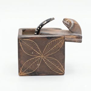 May include: A dark brown wooden box with a carved leaf design on the front. The lid is partially open, revealing a carved snake head and a black and white dotted handle. The box is likely used for storage or display.
