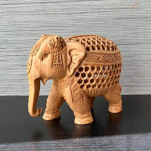 May include: A detailed wooden elephant sculpture with intricate carvings. The elephant is light brown and features a decorative saddle and openwork design on its back. The sculpture is standing on a black surface.