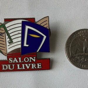 Op de afbeelding: Een rode, witte en blauwe emaille pin met een afbeelding van een open boek en de tekst "SALON DU LIVRE". De pin is ongeveer zo groot als een Amerikaanse kwart.