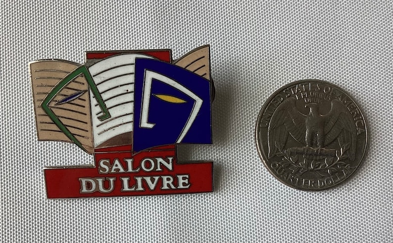 Op de afbeelding: Een rode, witte en blauwe emaille pin met een afbeelding van een open boek en de tekst "SALON DU LIVRE". De pin is ongeveer zo groot als een Amerikaanse kwart.