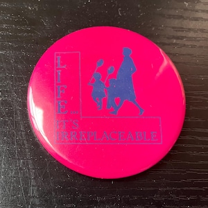 May include: A round, hot pink button with the words "LIFE... IT'S IRREPLACEABLE" in blue. A blue silhouette of a person and two children holding balloons is also featured on the button.