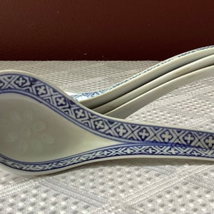 May include: A stack of three white ceramic soup spoons with a blue geometric border design. The spoons have a deep bowl shape and are stacked on top of each other. The spoons are on a white textured surface.