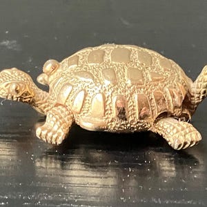 May include: A detailed, golden-colored turtle figurine. The turtle has a textured shell and detailed legs, head, and tail. The figurine is likely made of metal and is a decorative item.