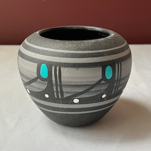May include: A small, round, gray ceramic pot with a black interior. The pot features horizontal gray stripes and decorative black and turquoise accents. The design includes black lines and white dots, creating a unique pattern. The pot is likely handmade.
