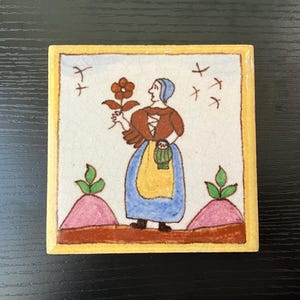 May include: A hand-painted ceramic tile featuring a woman in a blue dress and yellow apron holding a red flower. The tile has a yellow border and a white background with brown, pink, and green accents.