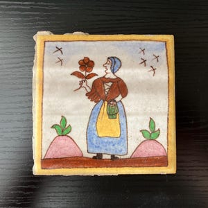 May include: A hand-painted ceramic tile featuring a woman in a blue dress and brown jacket holding a red flower. The tile has a yellow border and a white background with a blue sky and pink clouds.